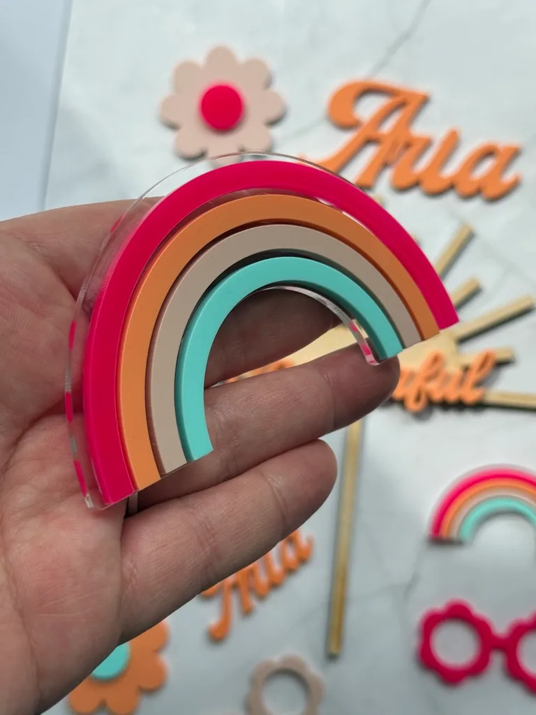 Layered acrylic rainbow held in a hand, in hot pink, orange, tan and mint